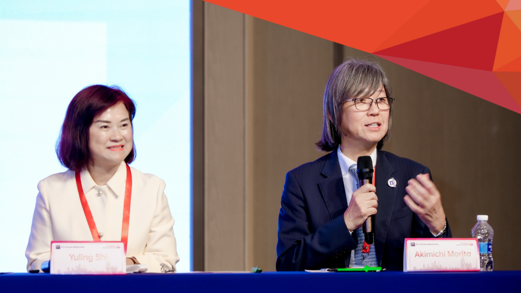 Two people sit at a table during a conference focused on regional outreach. The woman on the left smiles, while the man on the right speaks into a microphone. Nameplates in front identify them as Yuling Shi and Akimichi Morita.