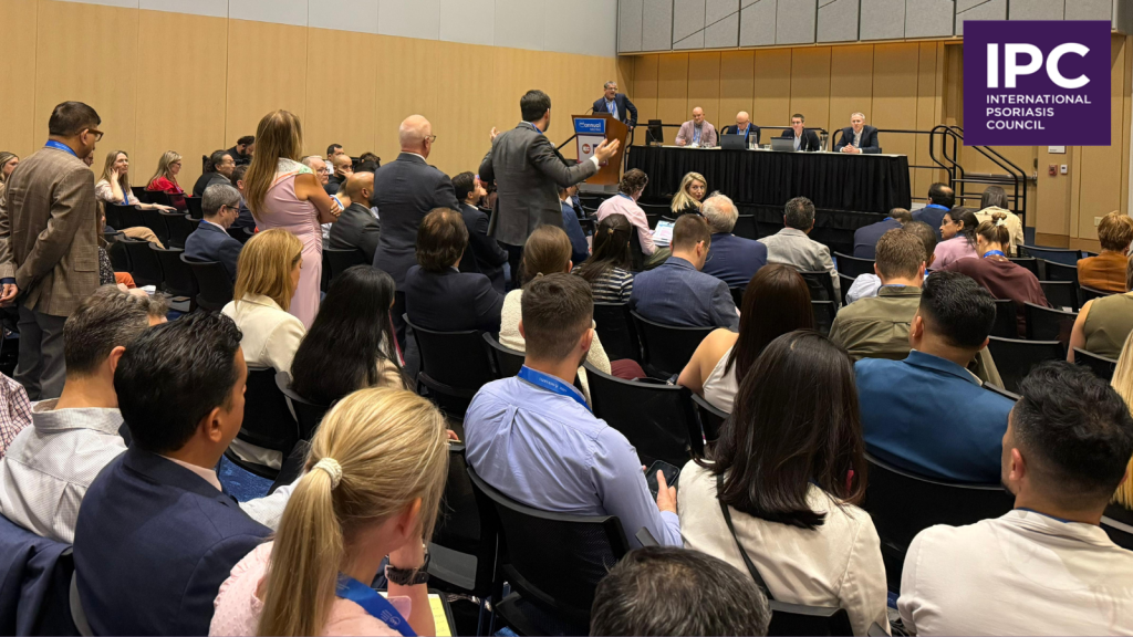 A large group attends a conference session, with several panelists seated at a table onstage. One attendee stands to ask a question. The IPC (International Psoriasis Council) logo and AAD 2026 appear in the corner.