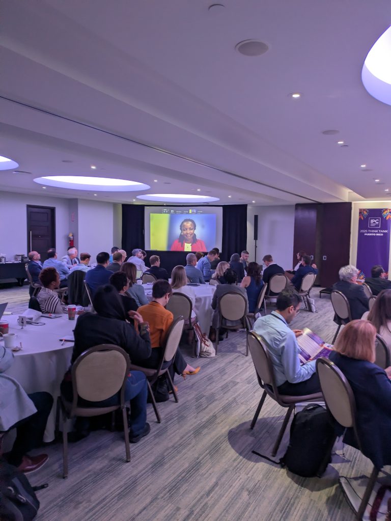 A conference room filled with people seated at round tables, watching a speaker on a screen at the front of the room. The atmosphere appears to be focused and attentive.
