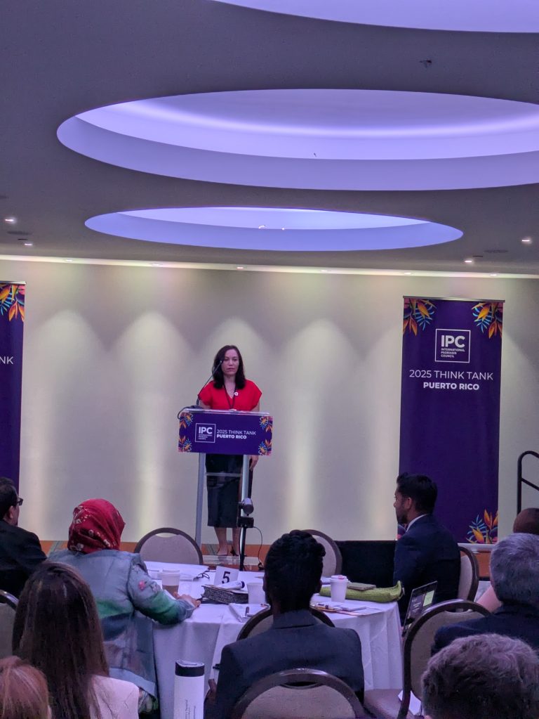 A woman stands at a podium speaking to an audience in a conference room. Two purple banners read IPC 2025 Think Tank Puerto Rico. People are seated at round tables, listening to the speaker.