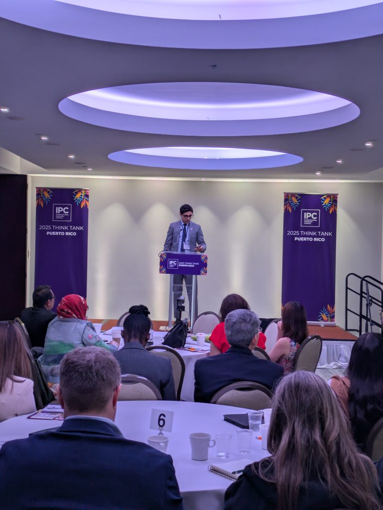 A man in a suit speaks at a podium on a stage, addressing an audience seated at round tables during the 2025 IPC Think Tank event in Puerto Rico. Two event banners are displayed behind him.