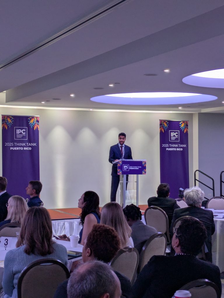 A man in a suit stands at a podium giving a speech at the IPC 2025 Think Tank event in Puerto Rico. Attendees sit at round tables listening. Colorful banners hang on either side of the podium.