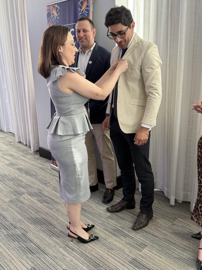 A woman in a gray dress pins a badge on a man in a beige blazer while another man stands nearby, all smiling in a well-lit room with white curtains and patterned carpet.