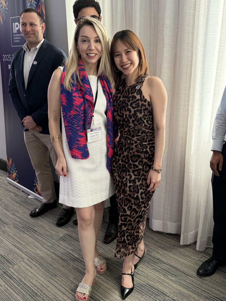 Two women pose and smile at an indoor event; one wears a white dress with a colorful scarf, the other a sleeveless leopard print dress. Several people stand in the background near a conference banner.
