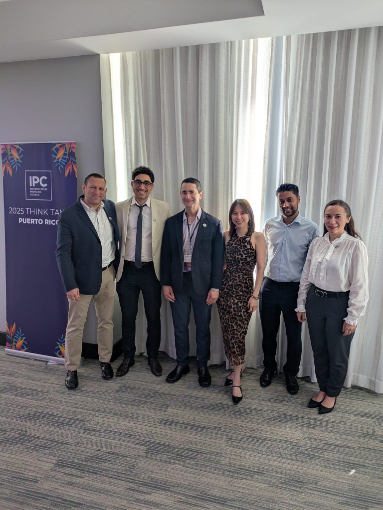 Six people stand in a row, smiling for a group photo indoors near a window with white curtains. A sign on the left reads “IPC 2025 Think Tank Puerto Rico.” The group is dressed in business or business-casual attire.