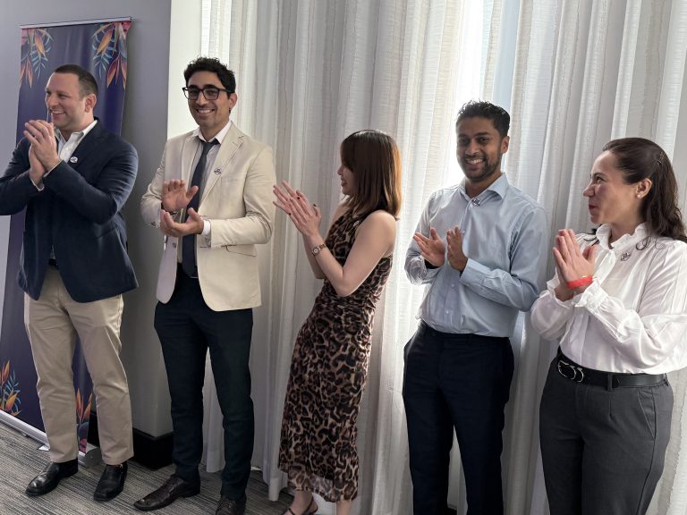 Five people stand in a row indoors, smiling and clapping. They are dressed in business or semi-formal attire. Light streams in through sheer curtains behind them, creating a bright, cheerful atmosphere.