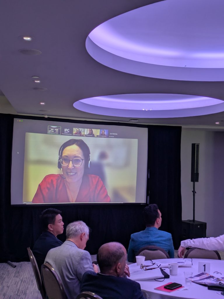 A woman wearing glasses and a headset speaks on a large screen during a video call at a conference. Several people sit at round tables in a dimly lit room, watching the presentation.